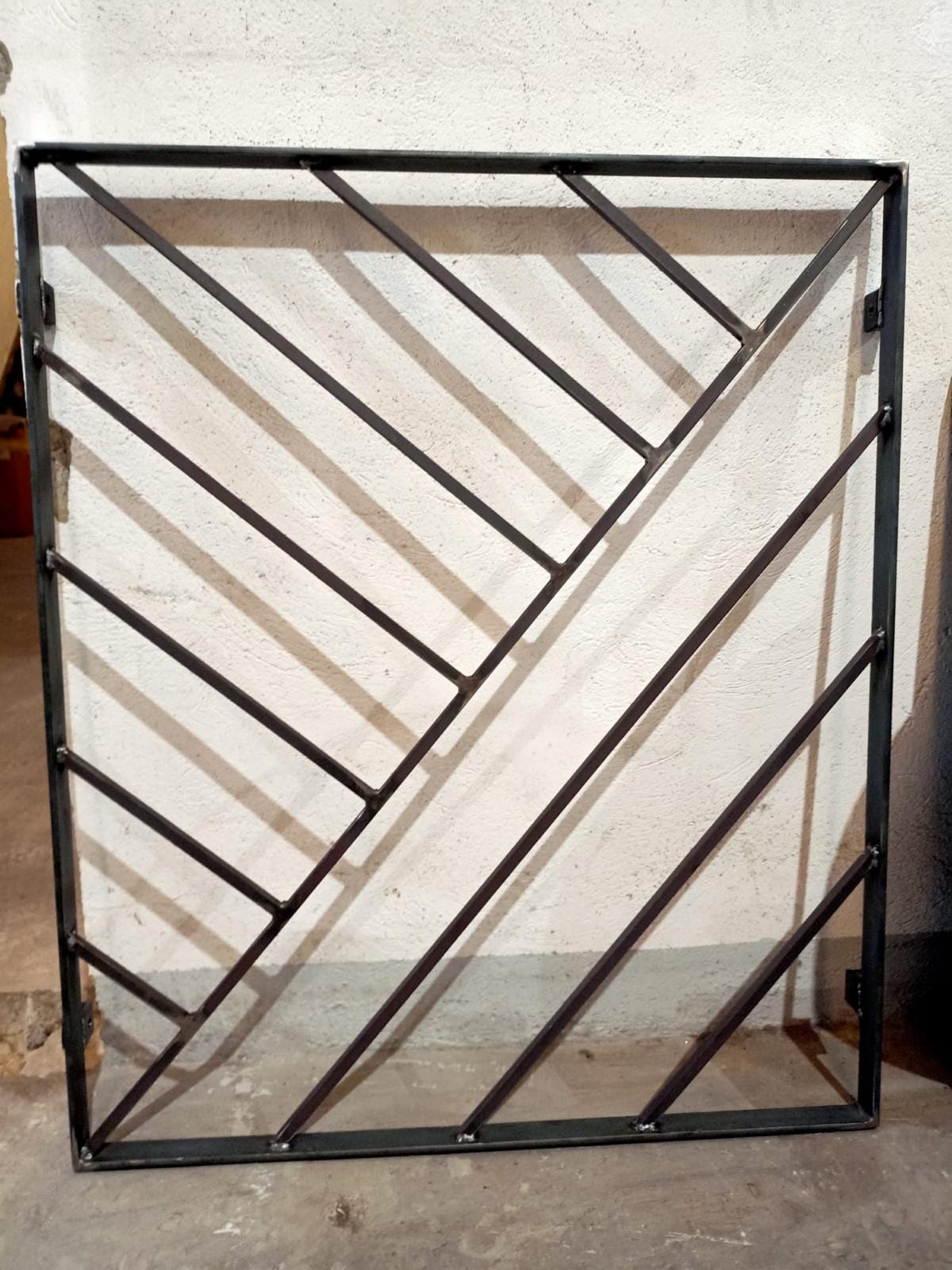 Reja recién soldada en acero sin pintar, con marco de tubo cuadrado, diagonales en espiga y pletinas de anclaje, apoyada contra una pared del taller.