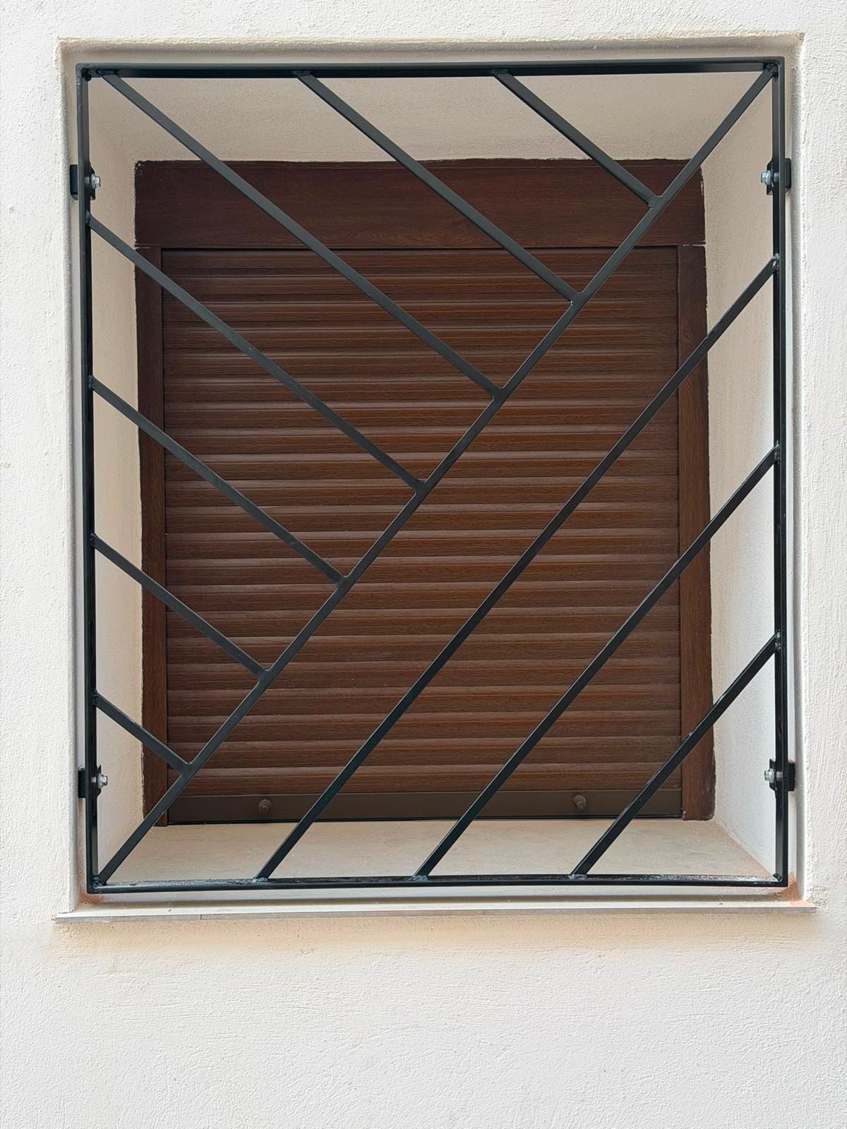 Reja negra de diagonales en espiga instalada en una ventana de fachada blanca, con la persiana de madera bajada detrás.